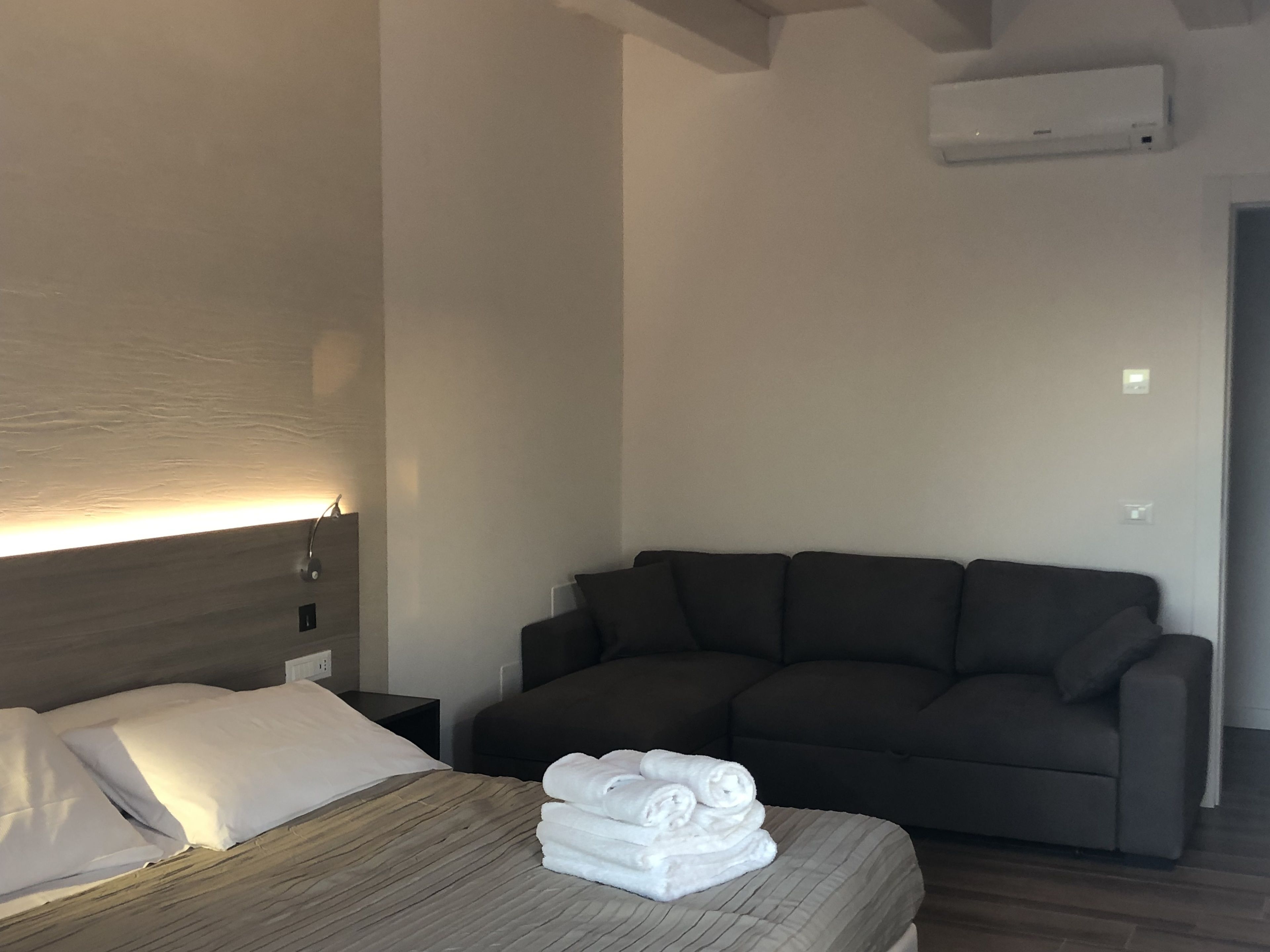 Habitación de hotel con cama, sofá oscuro y una pila de toallas blancas sobre la cama.