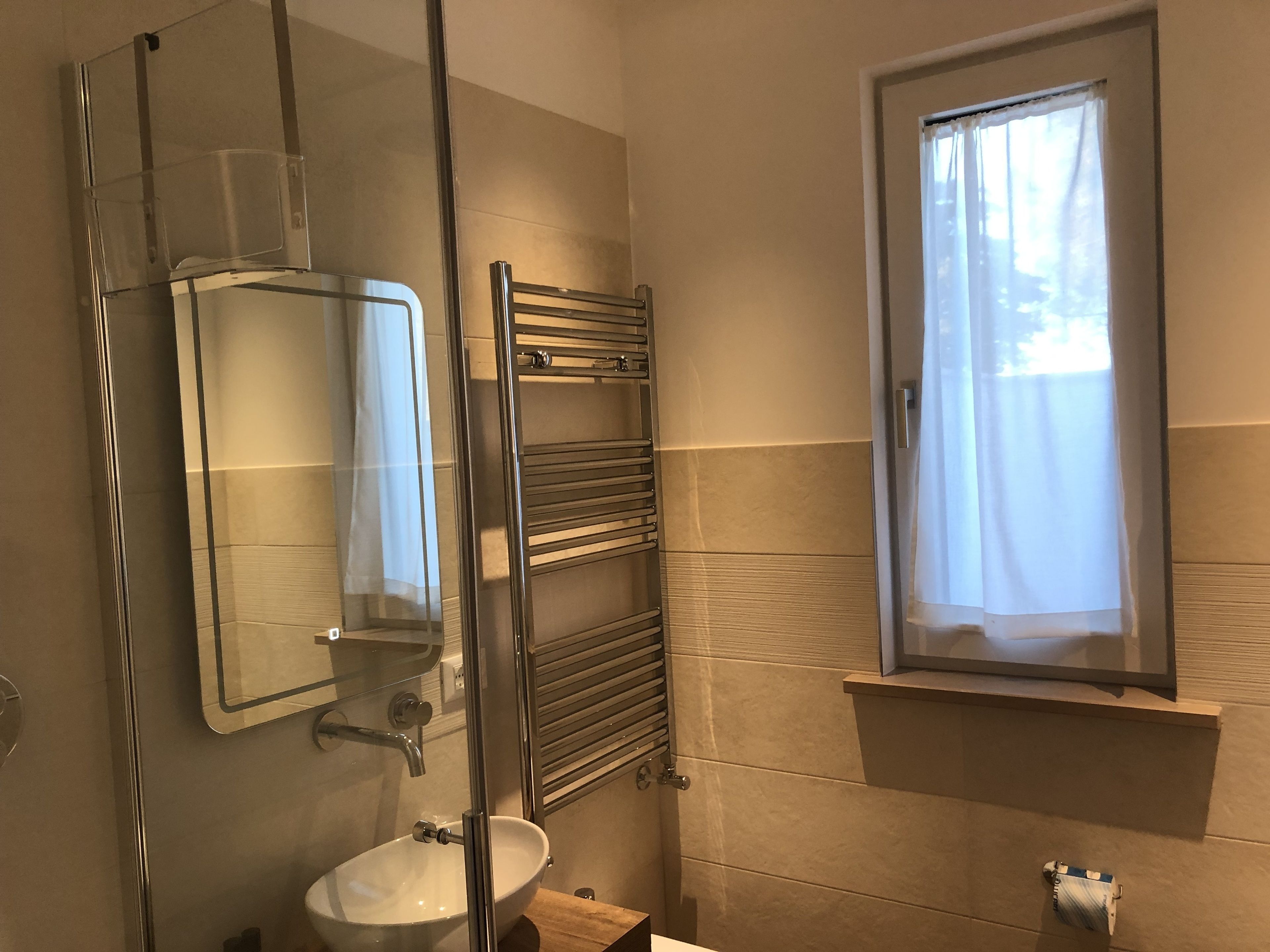 A bathroom with a glass shower enclosure, a sink, a towel rack, and a window with curtains.