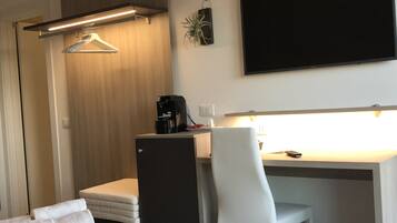 Habitación de hotel con cama, escritorio con silla, cafetera, televisor y planta.