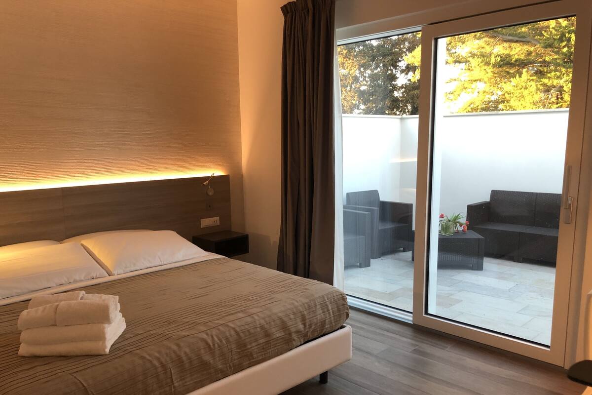 Una habitación de hotel moderna con una cama grande, una mesita de noche, una lámpara de pared y un ventanal corredizo que da a una zona de estar exterior.