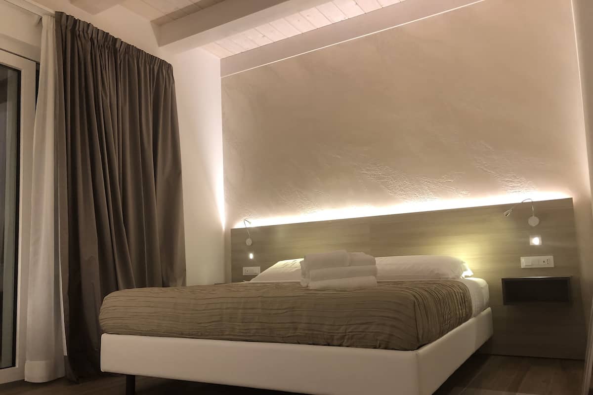 Un dormitorio moderno con una cama grande, cabecera con iluminación integrada, una mesita de noche y vistas al exterior a través de una ventana con cortinas.