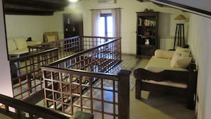 A spacious living room with a high ceiling, a wooden staircase, a sofa, a bookshelf, and a window with curtains.