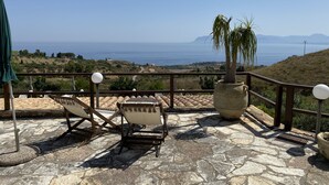 A terrace with stone flooring, two lounge chairs, a potted palm tree, and a view of the sea and hills.