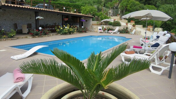 A swimming pool with lounge chairs and umbrellas, surrounded by potted plants and a stone building.