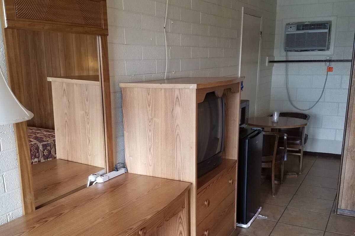 Quarto compacto com escrivaninha de madeira, televisão, mesa pequena e uma cadeira.