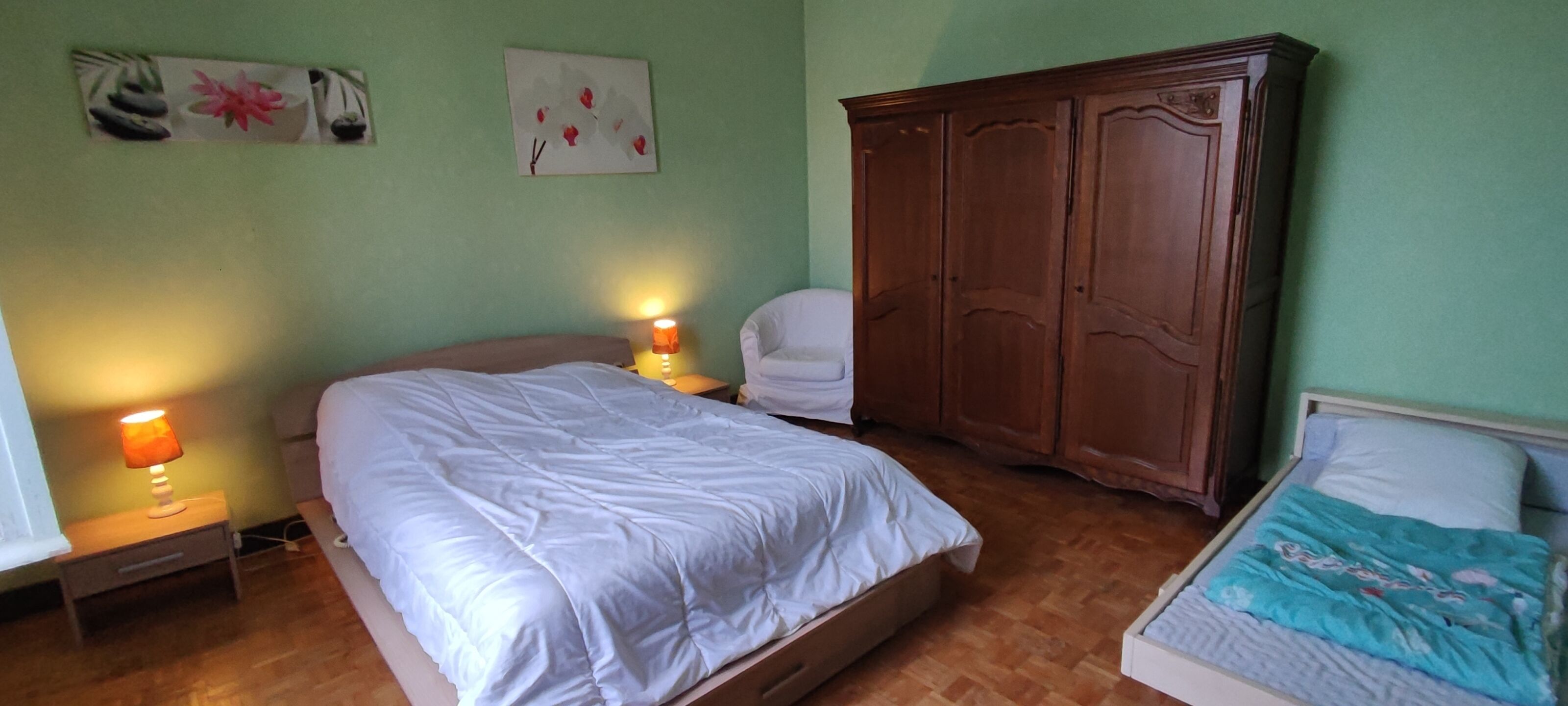 A bedroom with a large bed, a wardrobe, a nightstand with a lamp, and two wall-mounted paintings.