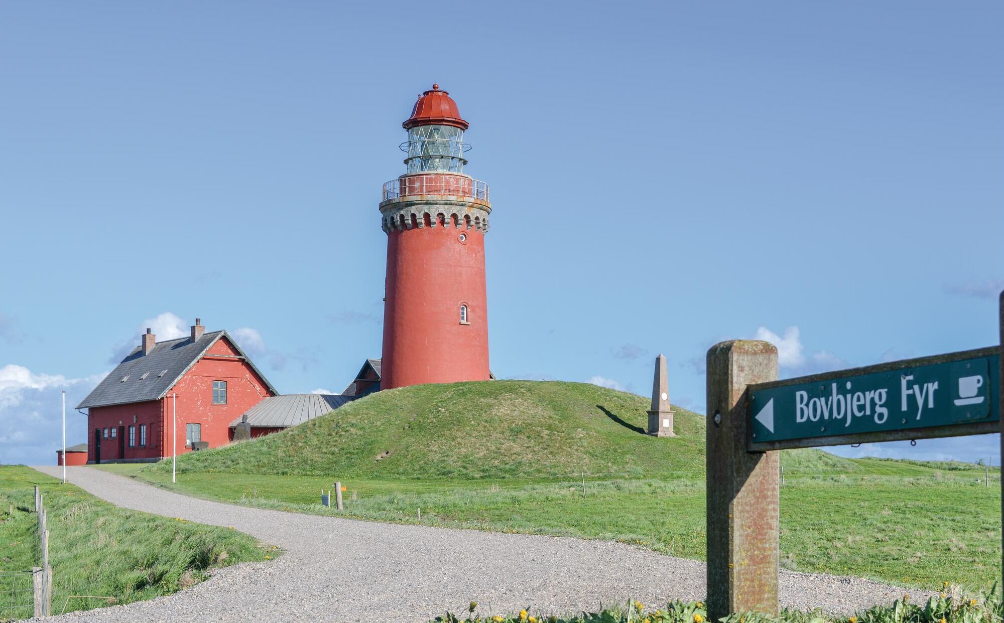 Un faro rosso con un sentiero ben definito che vi conduce, un edificio rosso nelle vicinanze e un cartello direzionale per 'Bovbjerg Fyr'.