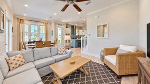A spacious living area with a sofa, a coffee table, and a wicker chair. There is a kitchen in the background with a dining area.