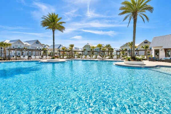 A large swimming pool with lounge chairs and palm trees, surrounded by multi-story buildings.