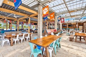 An outdoor dining area with wooden tables and chairs, banners hanging from the ceiling, and a view of the sky through a glass roof.