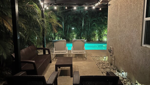 En udendørs terrasse med pool, fletmøbler, lyskæder og et pergola.