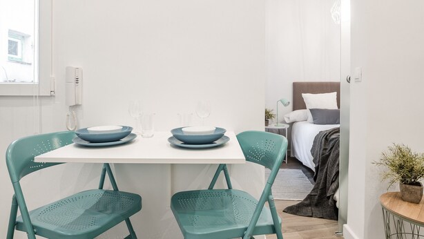 Une pièce de taille modeste, avec une table blanche et deux chaises de couleur turquoise, un lit recouvert d’une couette grise et blanche, et une petite plante en pot posée sur une table d’appoint en bois.