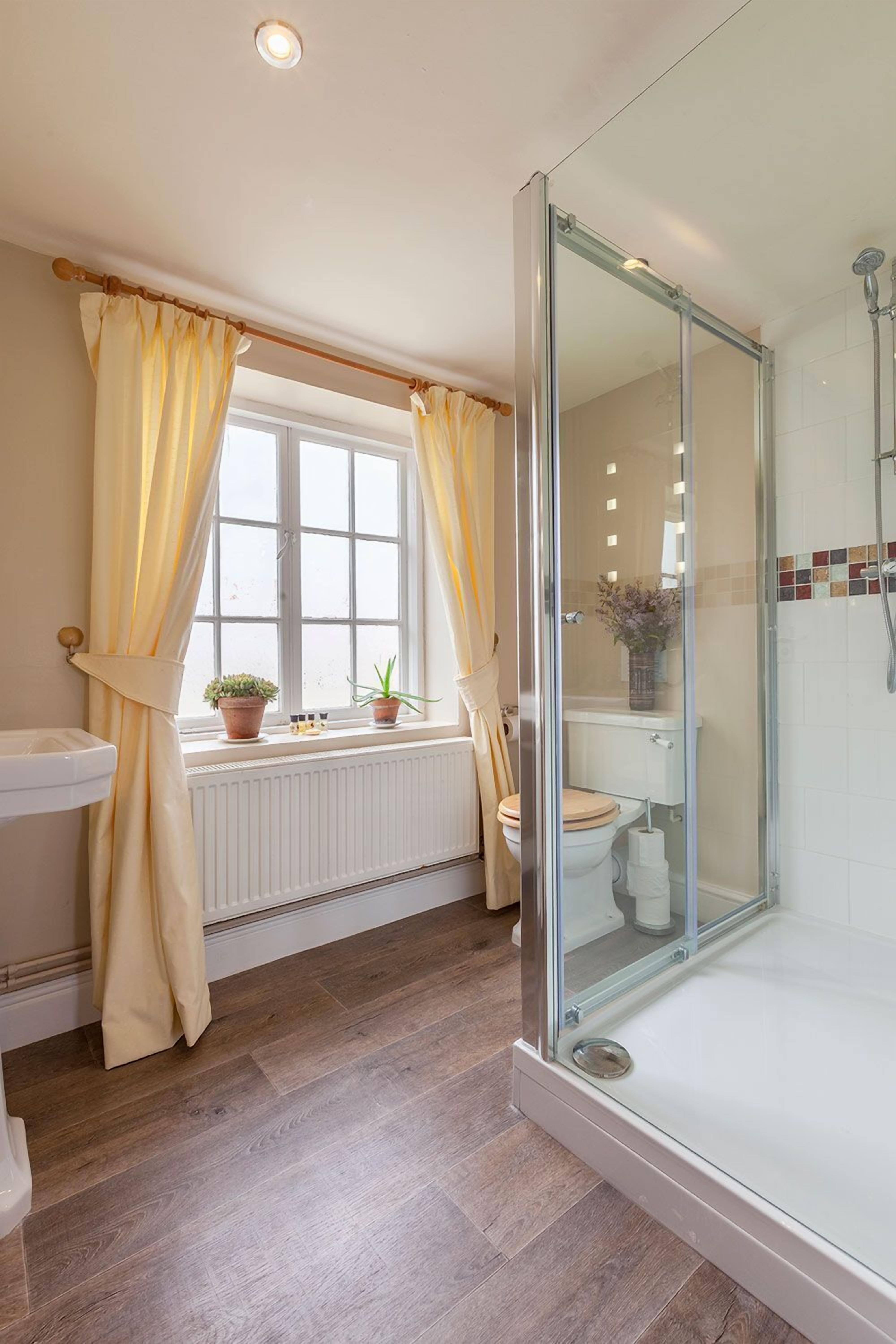 A bathroom with a glass-enclosed shower, a toilet, a sink, and a window with curtains.