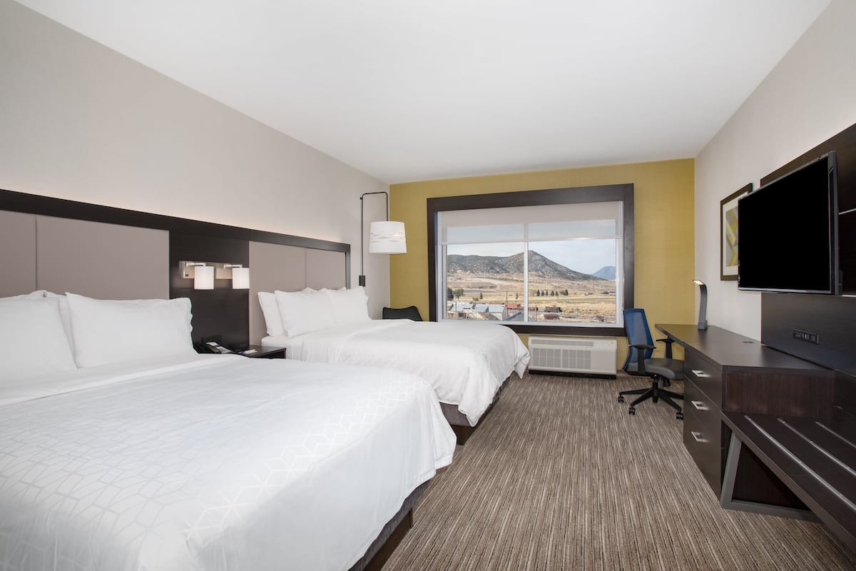 A hotel room with two beds, a desk, a TV, and a large window with a mountain view.