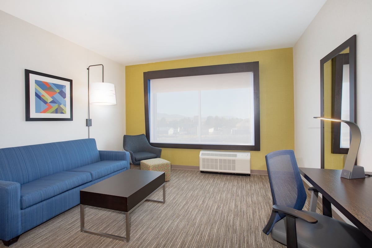 A hotel room with a blue sofa, a small table, a chair, a large window with a view, a framed picture, and a lamp.