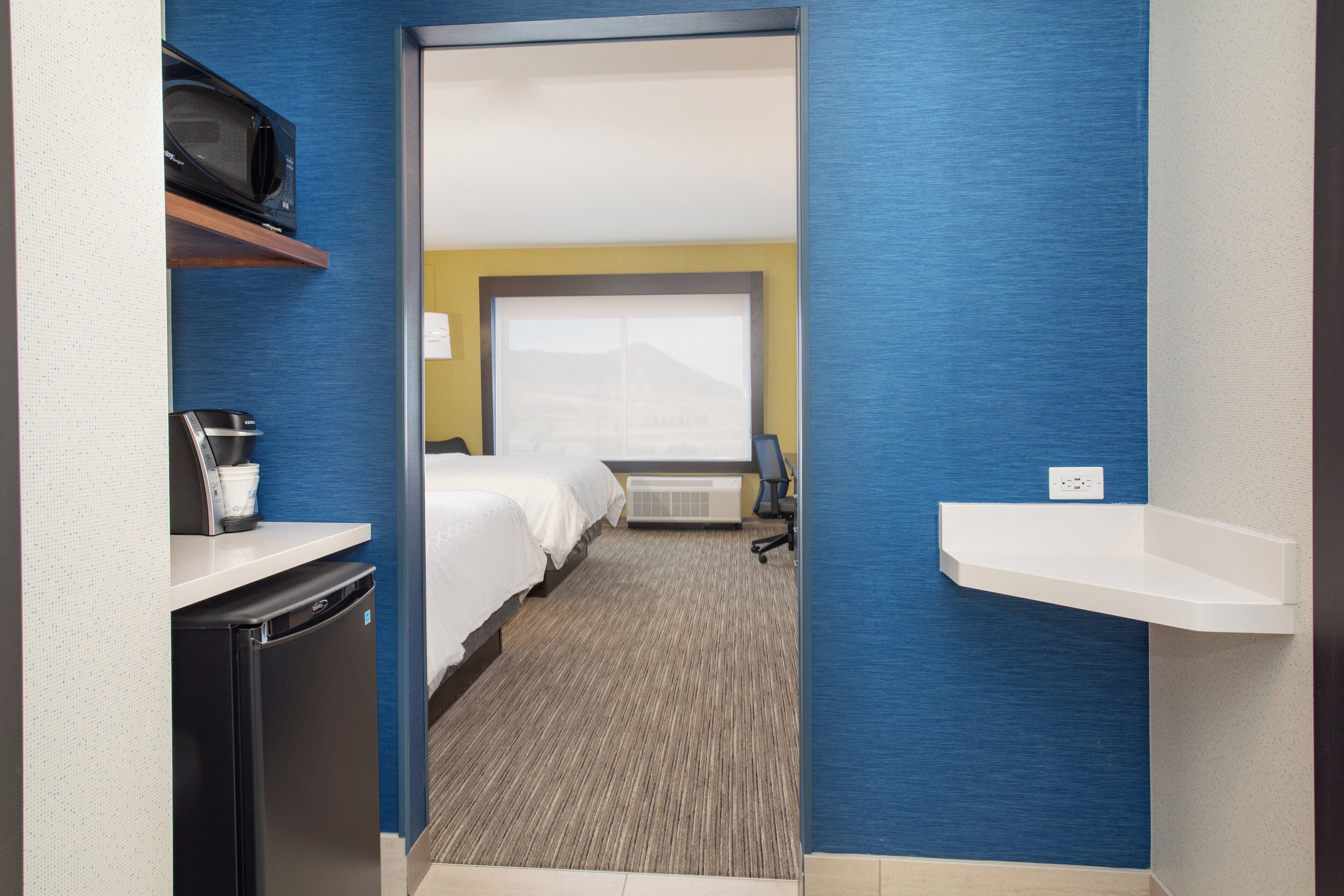 A hotel room with a bed, a desk with a coffee maker, a microwave, a window with a view, and a wall-mounted shelf.