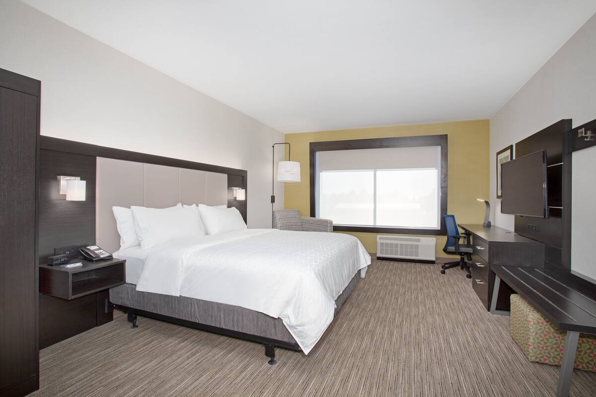 A hotel room with a bed, bedside table, lamp, desk, chair, and a large window.