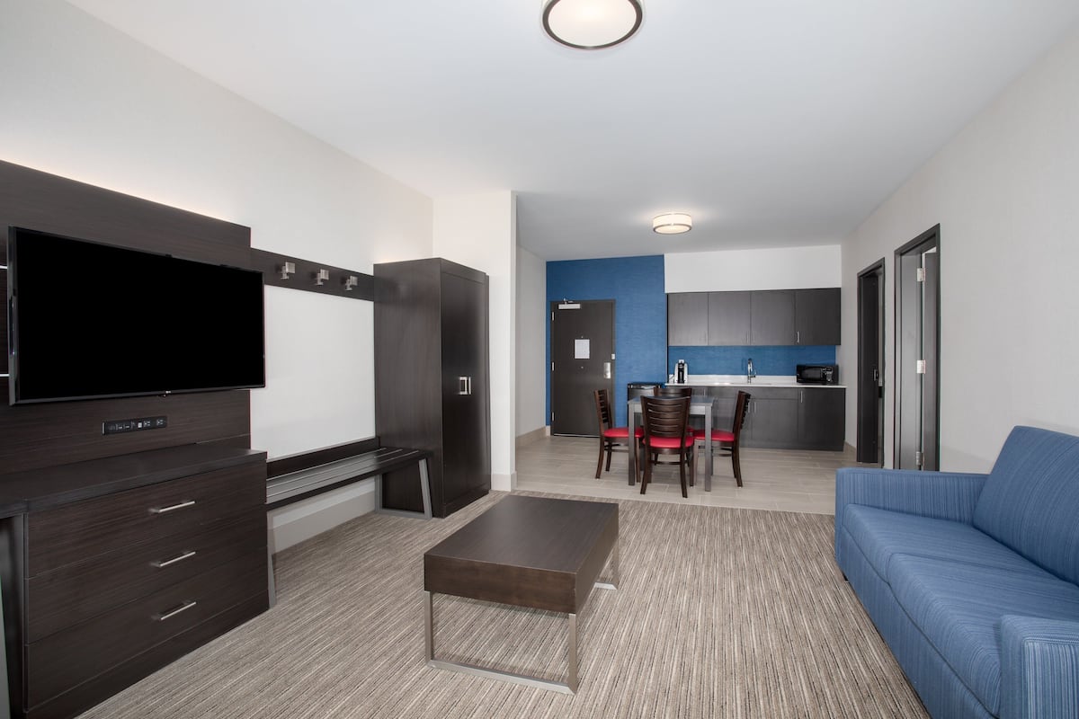 A modern living room with a flat-screen TV, dark wooden furniture, a blue sofa, and a kitchen area in the background.