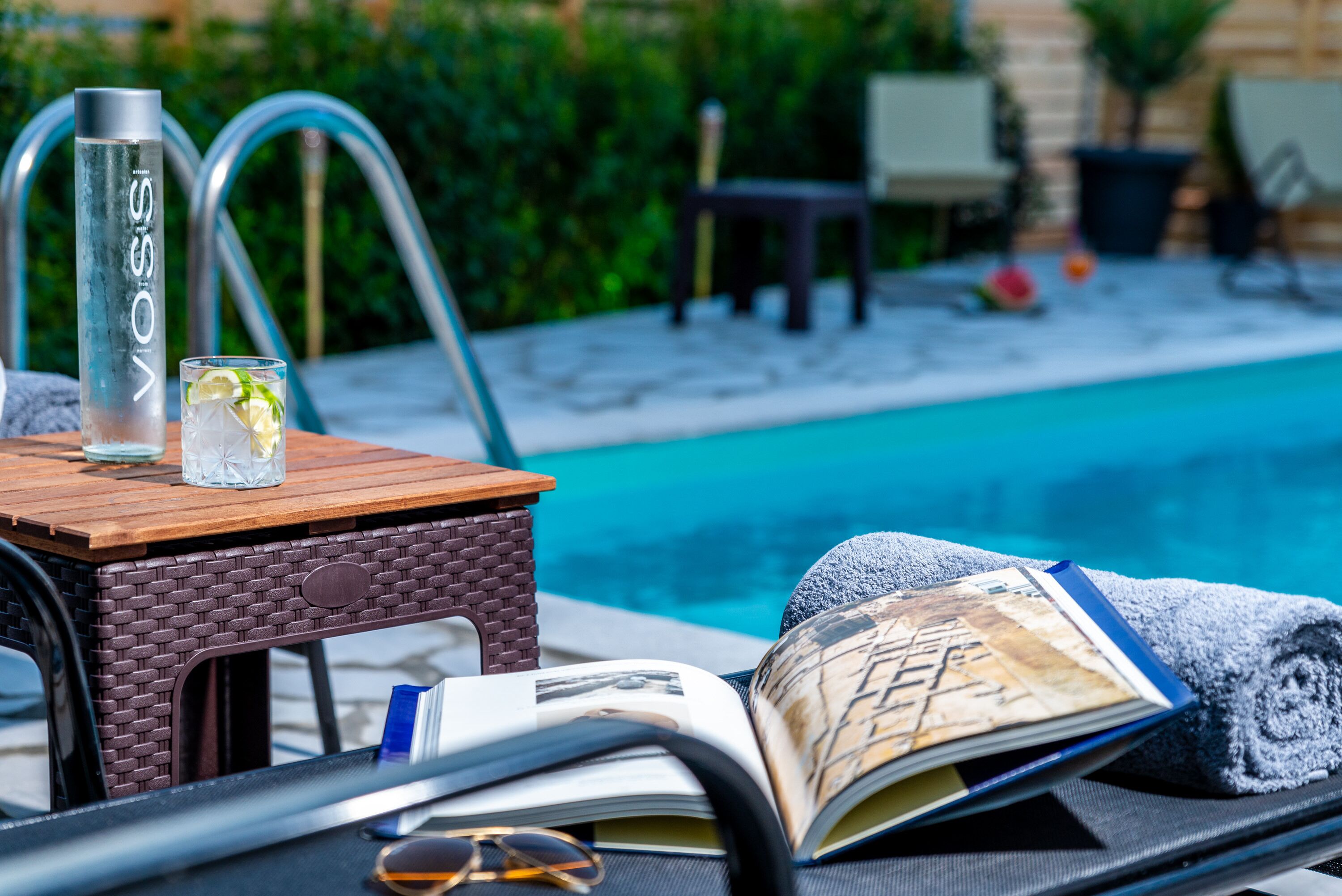 A poolside setting with a lounge chair, a book, a glass of water, and a bottle.