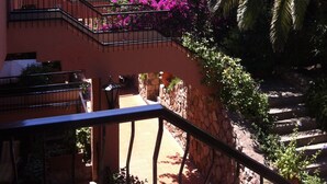 A balcony with a view of a lush garden, a stone wall, and a staircase leading to an upper level.