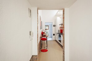 A narrow hallway leading to a kitchen with a red stool and a dining area in the background.