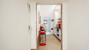 A narrow hallway leading to a kitchen with a red stool and a dining area in the background.