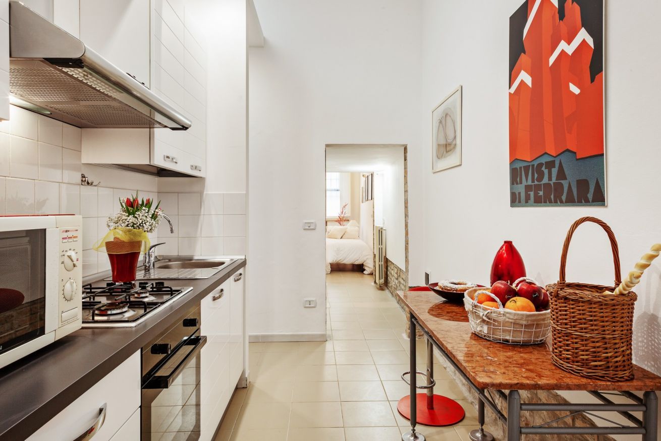 Cucina stretta con mobili bianchi, piano cottura a gas e un vaso rosso con fiori.
