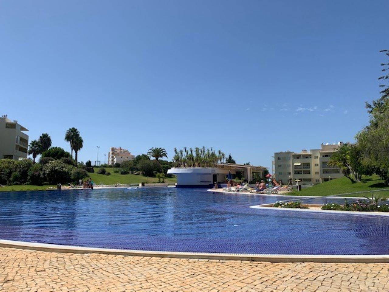 Ein großer Pool im Freien, mit blauem Himmel, umgeben von Grünflächen und Gebäuden.