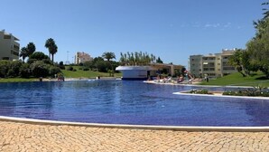 Une grande piscine extérieure, avec un ciel bleu dégagé, entourée de verdure et de bâtiments.