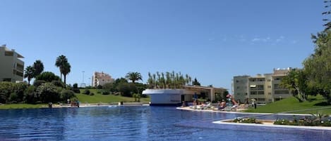 A large outdoor swimming pool with a clear blue sky, surrounded by greenery and buildings.