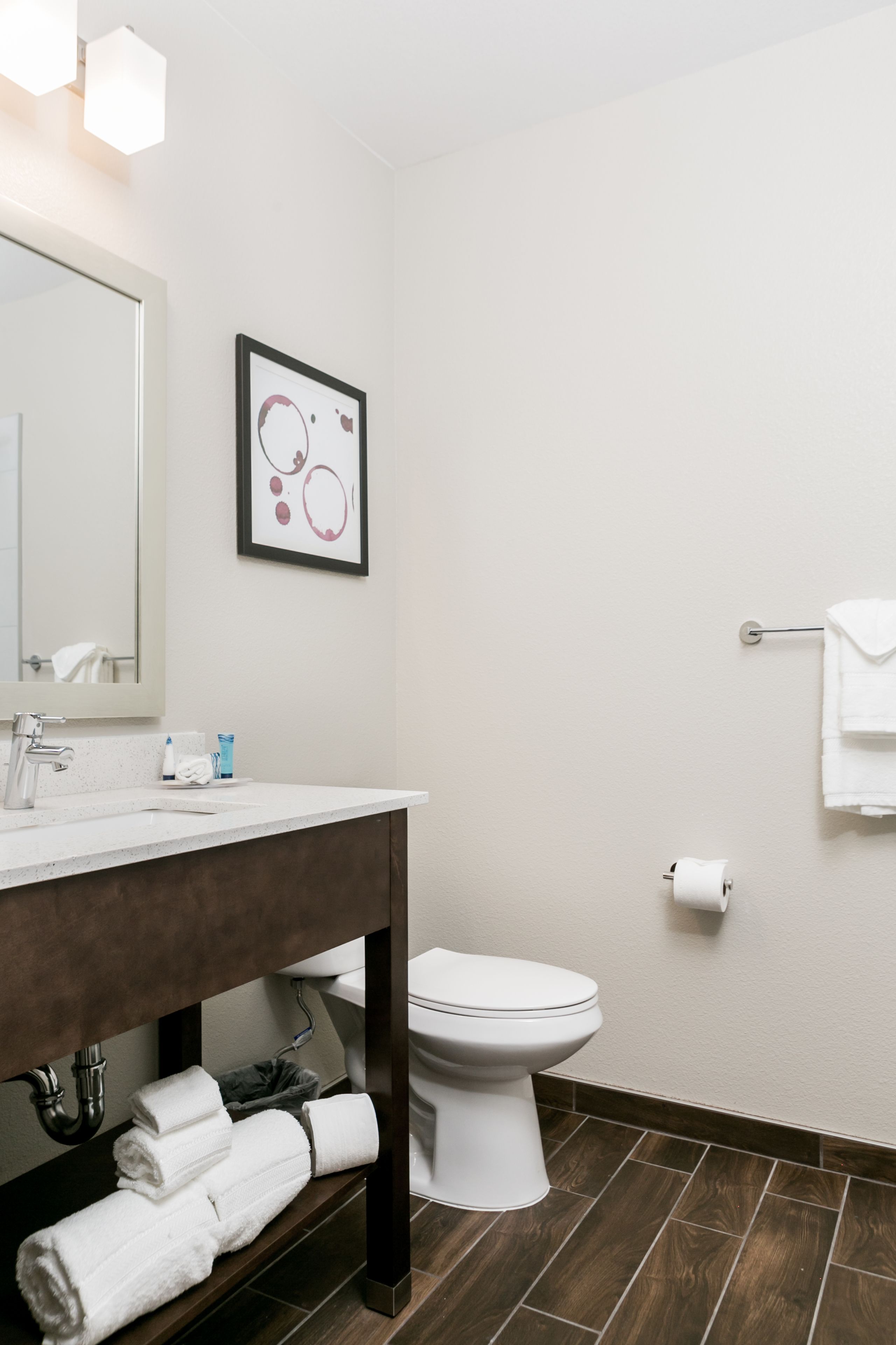 A modern bathroom with a white sink, mirror, and toilet. There is a framed artwork on the wall and a towel rack with a white towel.
