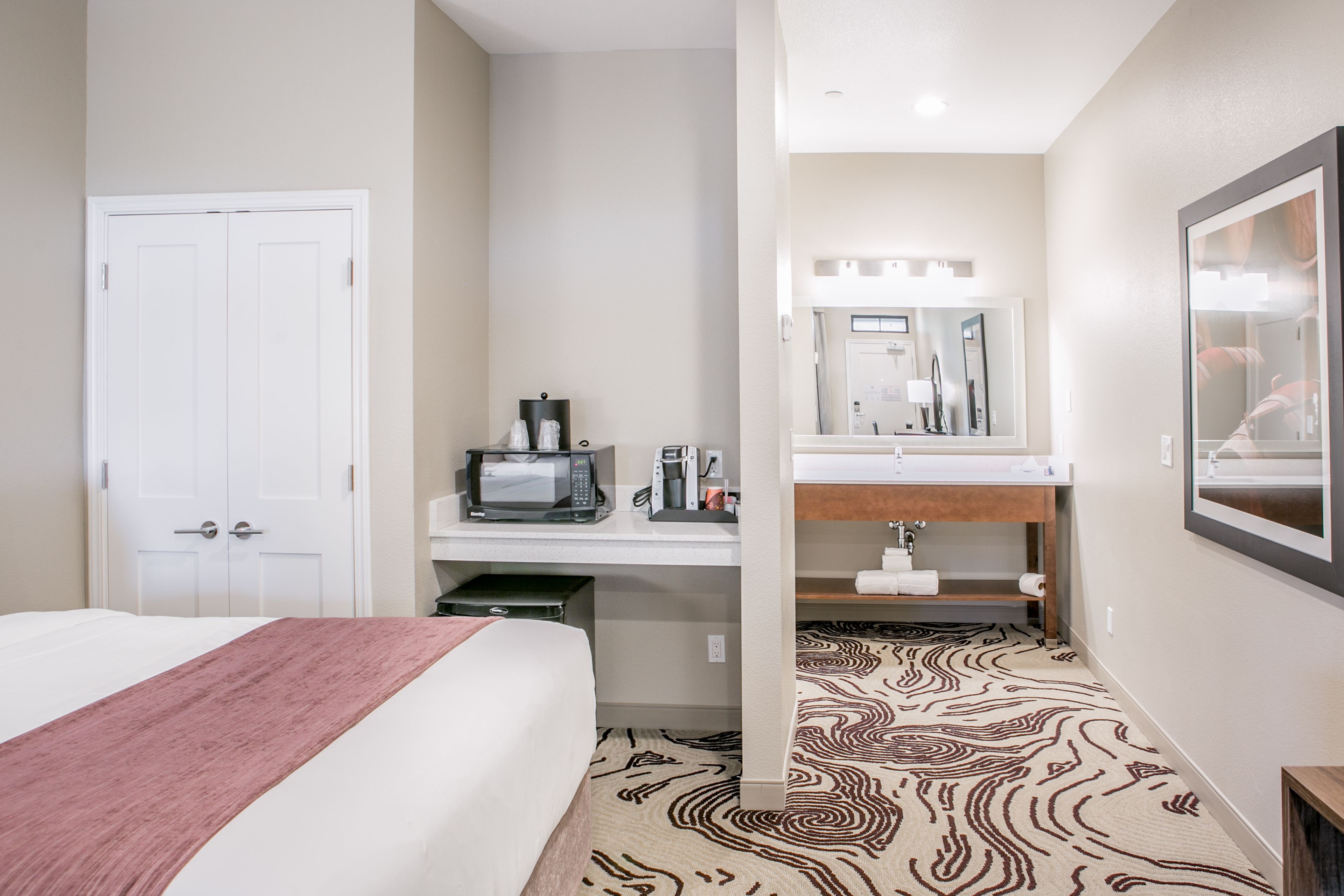 A hotel room with a bed, a desk with a microwave, a sink, and a mirror.