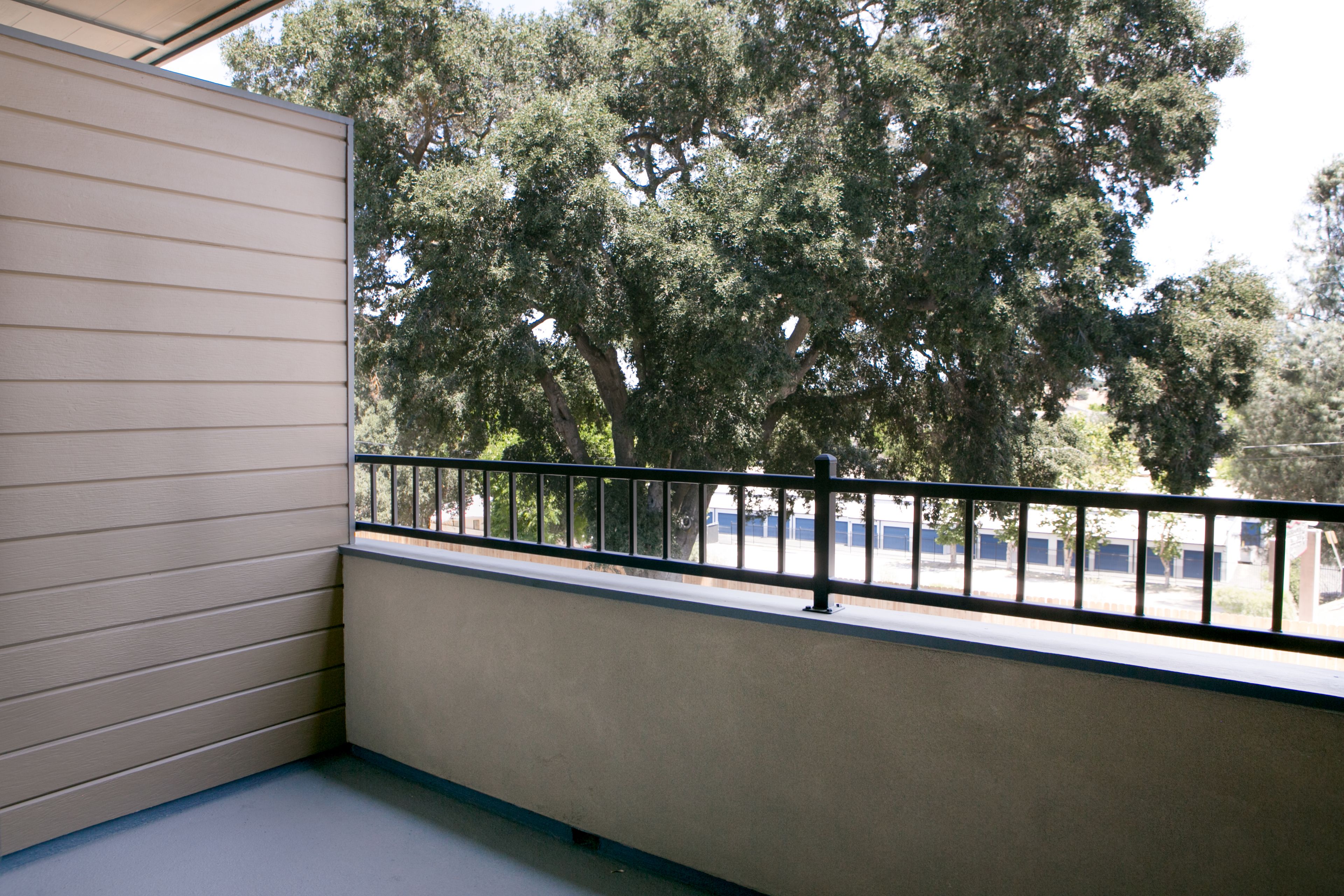 A balcony with a metal railing, beige walls, and a view of trees and a parking lot.