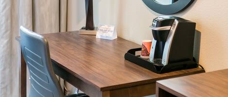 A hotel room with a wooden desk, a black chair, a coffee machine, a lamp, and a round mirror on the wall.
