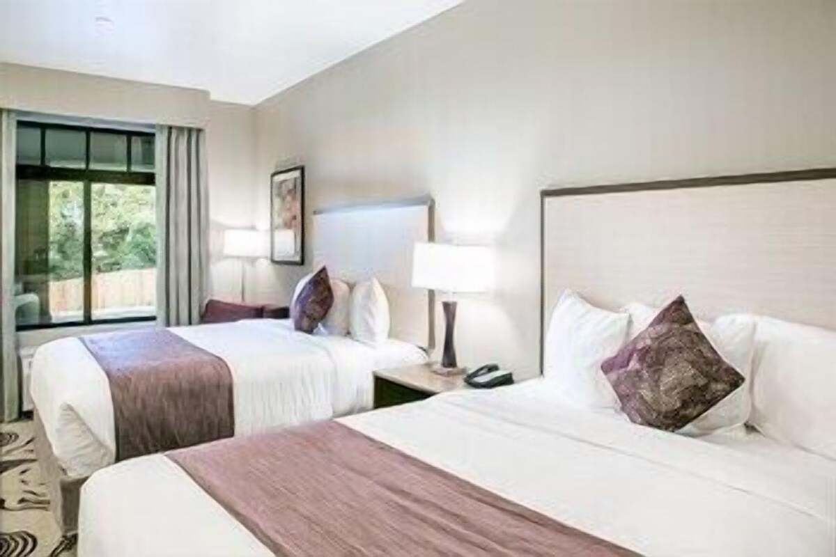 A hotel room with two beds, a large window, a bedside table with a lamp, and a framed picture on the wall.