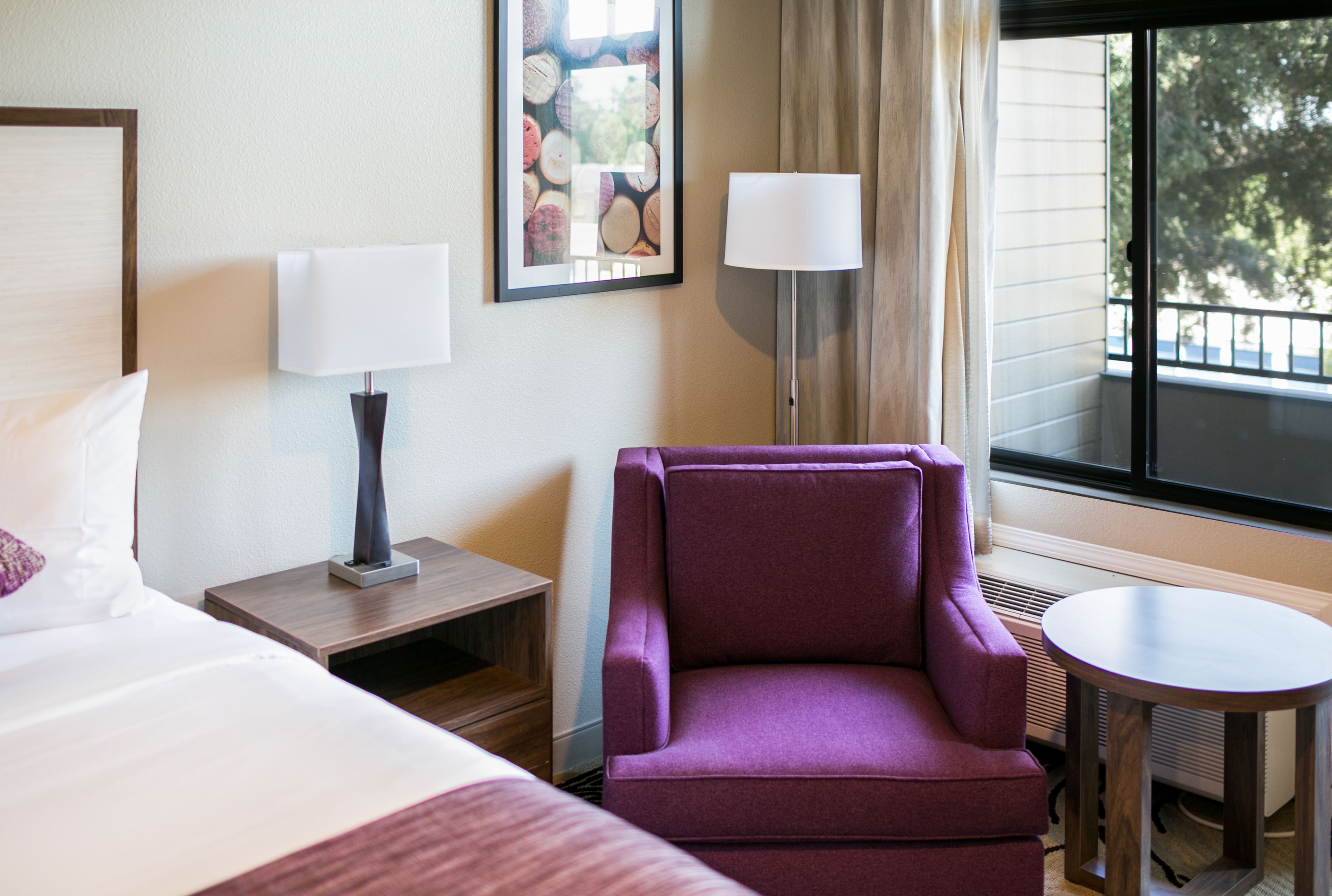 Quarto de hotel com uma cama, uma poltrona roxa, uma mesinha, uma luminária e janela com vista para árvores.