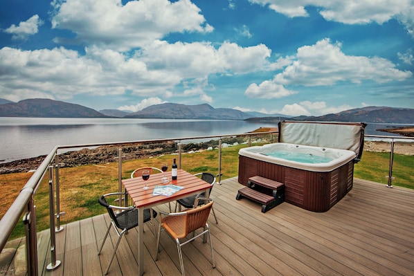 A deck with a hot tub, table, and chairs overlooking a lake with mountains in the distance.