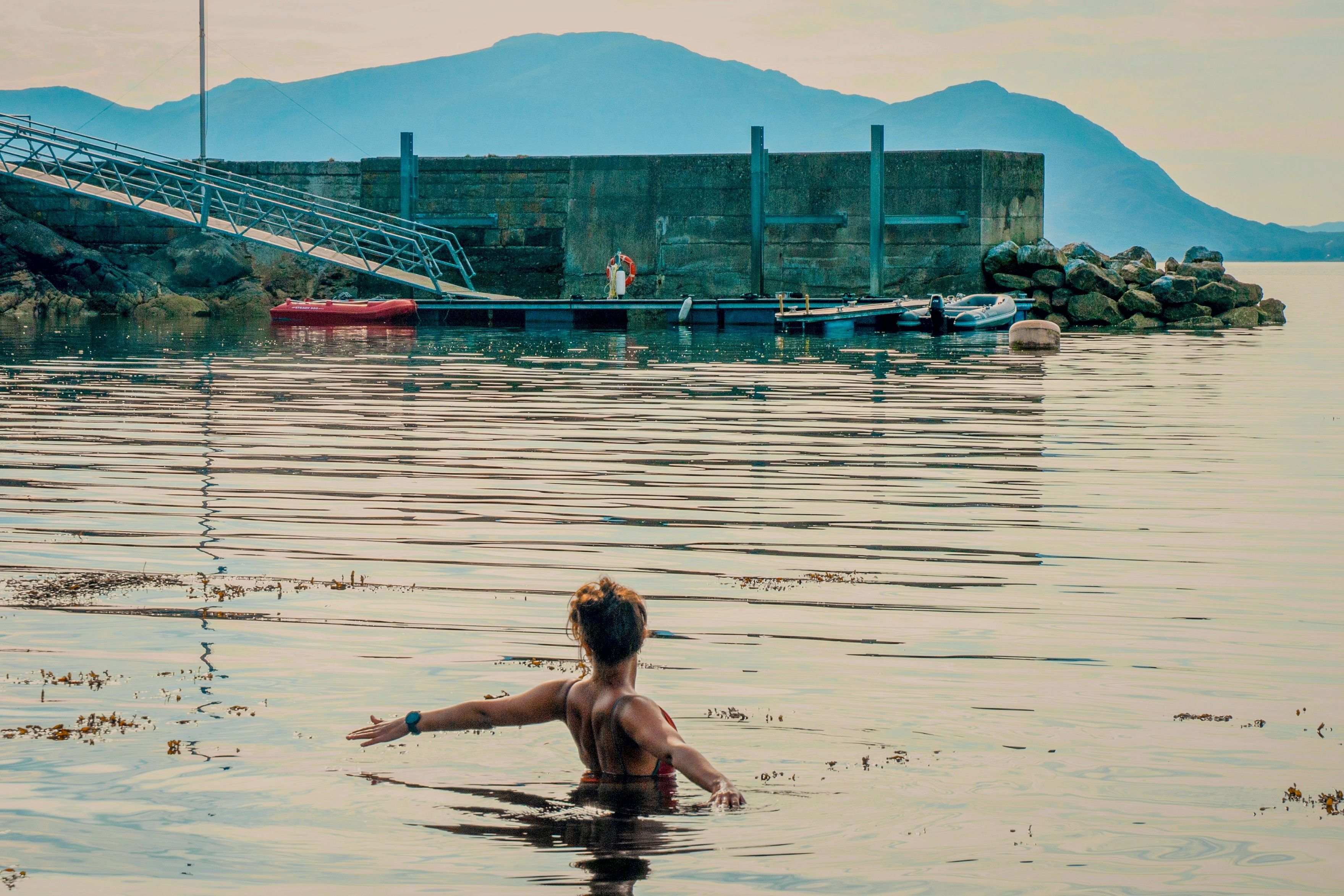 Una persona en el agua, con un muelle y barcos al fondo.