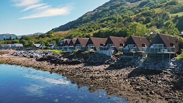A row of chalet-style cabins with steep roofs, situated on a rocky shoreline with a backdrop of hills and a clear sky.