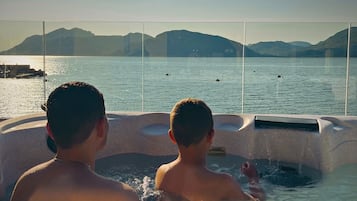 Two individuals in a hot tub with a view of a calm body of water and distant mountains.