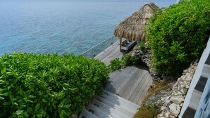 Une terrasse en bois surmontée d’une structure à toit de chaume, entourée d’une végétation luxuriante et offrant une vue sur une mer calme.