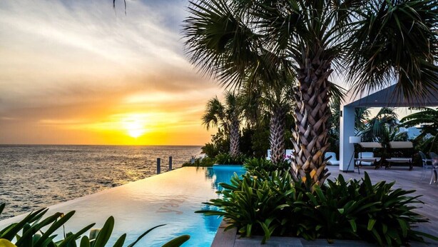 Une piscine à débordement offrant une vue imprenable sur le coucher de soleil sur l’océan, entourée de palmiers et agrémentée d’un espace salon extérieur moderne.
