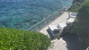 Une terrasse en bois avec des meubles d’extérieur blancs, donnant sur une mer d’un bleu limpide.