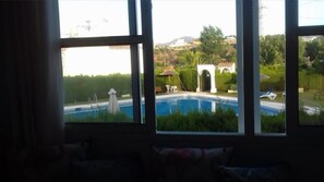 Blick durch große Fenster auf einen Swimmingpool, Gartenmöbel und ein weißes Gebäude mit Bögen.
