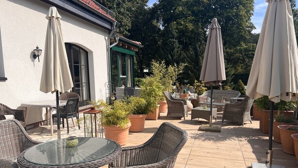 An outdoor patio area with wicker furniture, potted plants, and umbrellas. - Time by the Lake. Wildrose Duplex – Fireplace, Loggia, View of the Greenery (Bad Saarow)