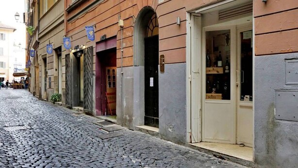 Uma rua de paralelepípedos com edifícios que apresentam portas em arco e uma loja com uma placa que diz 'MASI'.