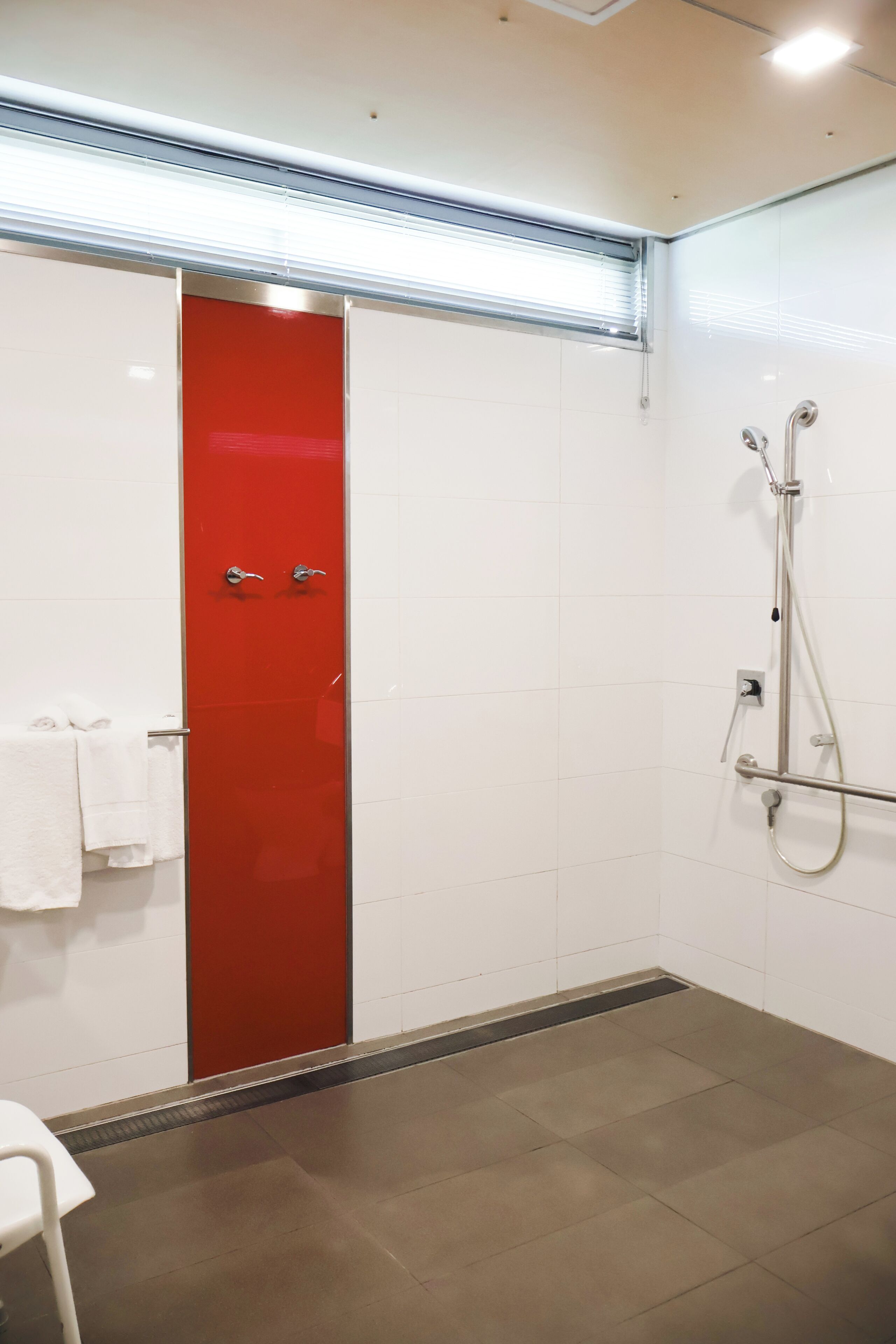 A bathroom with a red door, white tiles, a shower area, and a white chair.