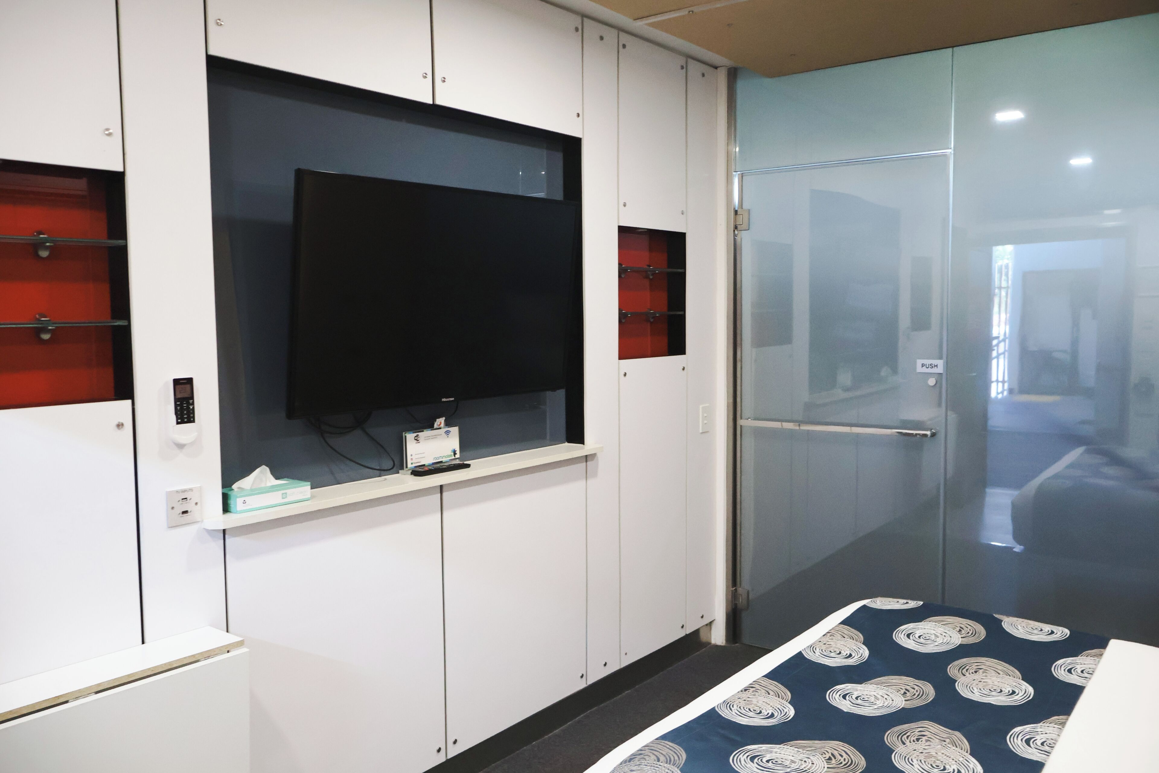 A hotel room with a television mounted on a white cabinet, a glass door leading to a hallway, and a patterned bedspread.