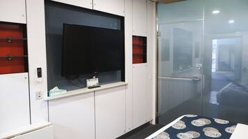 A hotel room with a television mounted on a white cabinet, a glass door leading to a hallway, and a patterned bedspread.