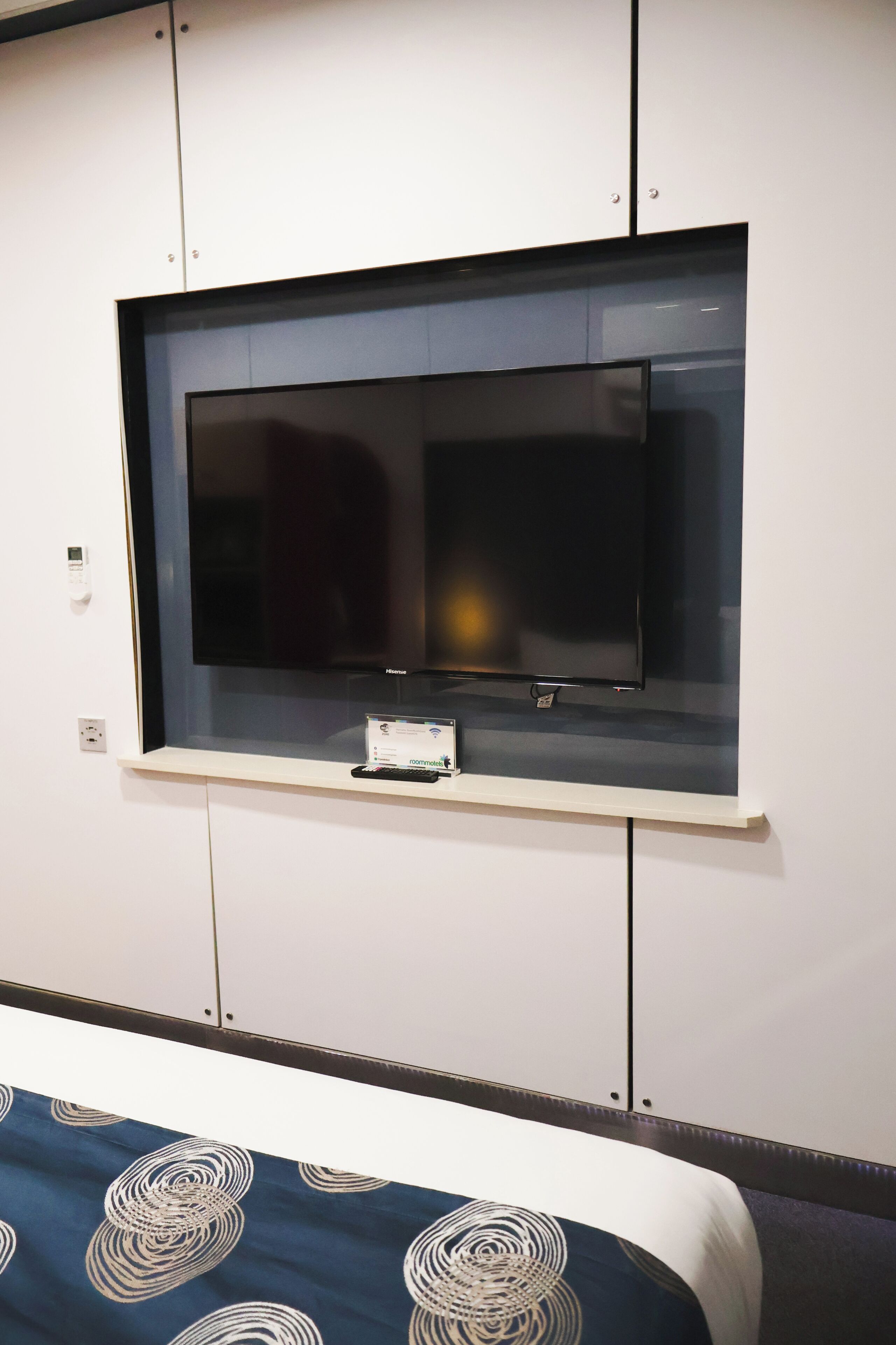 A hotel room with a television mounted on a wall, a bed with a patterned bedspread, and a small table with a remote control.